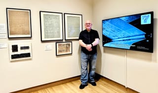 Glenn Fleishman standing between framed newspaper flongs (molds) and plates and an original and a video screen showing his video playing with captions on