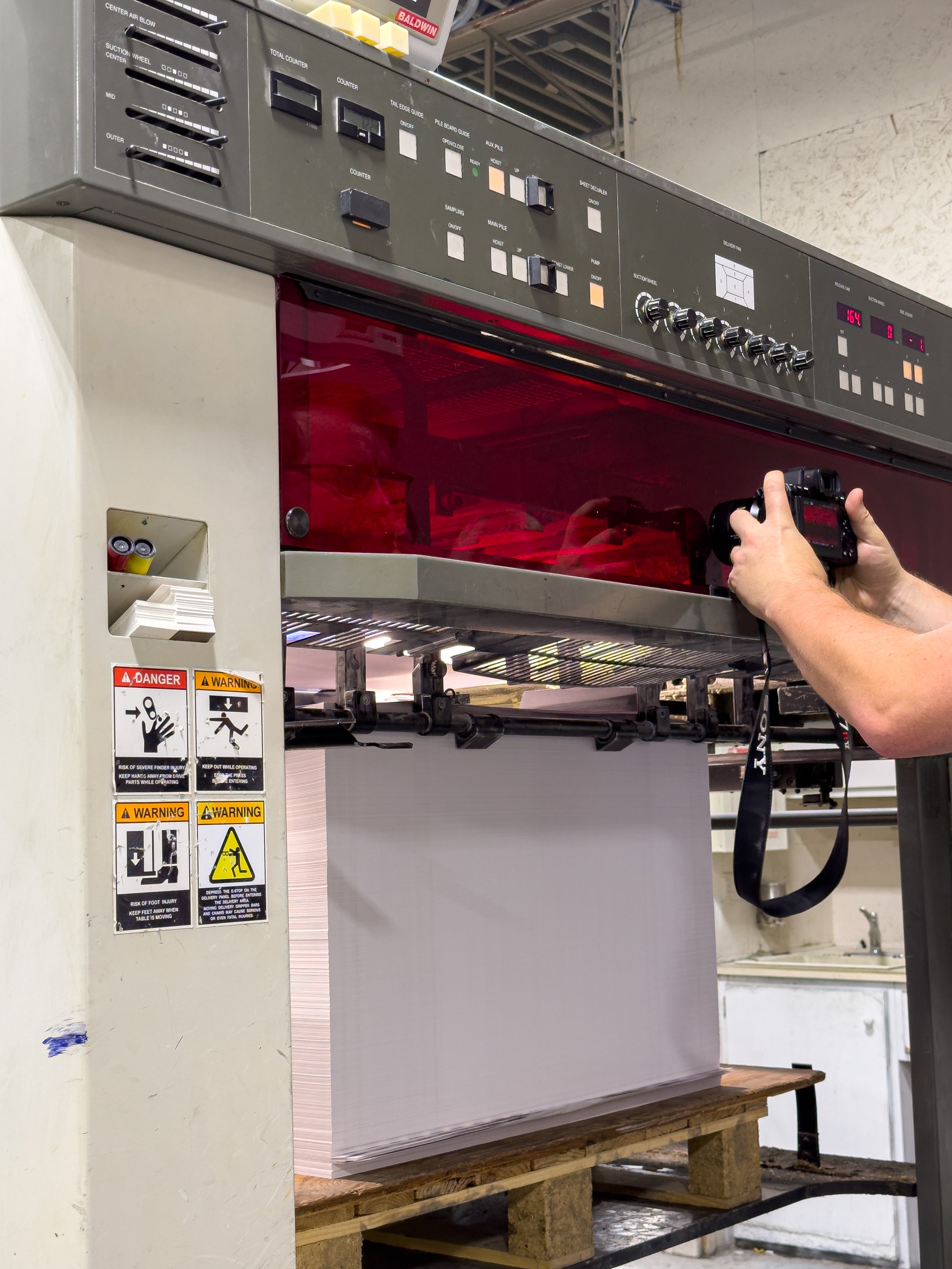 At the finished end of the press, a stack of paper grows as a ratcheting platform lowers, so the paper drops onto about the same height each time. A UV unit and a heater cure and then dry the ink instantly as the page passing through before landing on the stack.