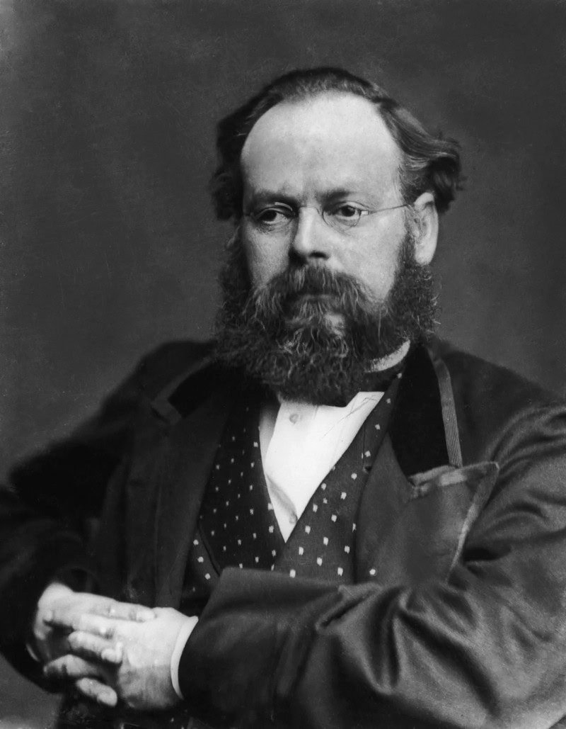 Photo of man in small glosses with mid-length hair and heavy beard, wearing a dotted vest, collarless shirt, and bowtie, in a formal jacket, with his hands holding each other